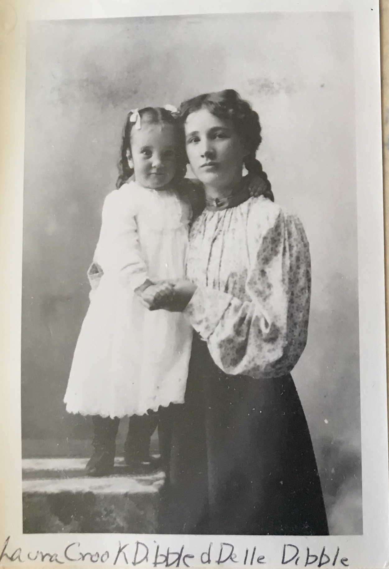 IMAGE/PHOTO: Laura Crook Dibble and daughter Della Dibble: Black and white photo of Della Dibble, about age 3, wearing a calf-length white dress and hair ribbons,standing on a table and holding the hand of her mother, Laura Dibble, wearing a loose-fitting white patterned blouse and dark skirt, with her braided hair falling down behind her back, standing beside her